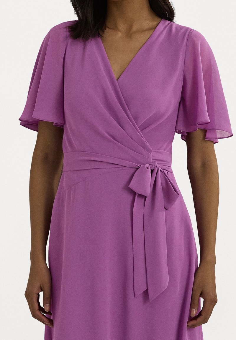 Šaty s výstřihem do V z jemné látky v barvě levandule, s krátkými volánkovými rukávy a detaily v oblasti pasu s uvázaným opaskem. Sukně je splývavá.