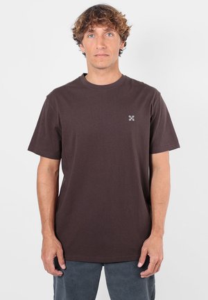 Homme aux cheveux bouclés portant un t-shirt marron foncé uni à manches courtes avec un petit logo blanc sur la poitrine gauche, debout devant un fond blanc.