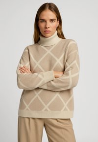 Maglione beige a collo alto con motivo a diamante diagonale color crema, texture morbida, colletto e polsini a coste, e vestibilità comoda.
