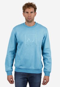 Sudadera azul claro de algodón, con un logo cuadrado que tiene las iniciales bordadas "AJ" en el centro. Textura suave, cuello redondo.