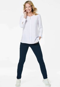 Blusa bianca a maniche lunghe con dettaglio a chiave, abbinata a pantaloni blu scuro e sneaker bianche, in piedi su uno sfondo neutro.