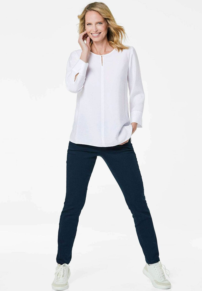 Witte blouse met lange mouwen en sleutelgatdetail, gecombineerd met donkerblauwe broek en witte sneakers, staande tegen een neutrale achtergrond.