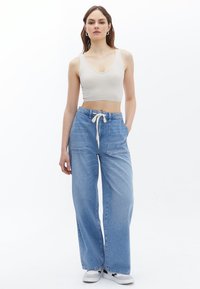 Ljusblå vida jeans med två framfickor och dragsko i midjan, kombinerat med en beige ribbad croptop. Modellen står mot en vit bakgrund.