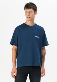 Navy blue cotton t-shirt with short sleeves and a round neck. Features a small white logo on the left chest. Straight cut and no patterns.