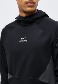 Άνδρας που φοράει μαύρο φούτερ Nike Pro με λευκό λογότυπο στο στήθος, εμφανίζεται από τους ώμους μέχρι τα χείλη σε απλό φόντο.