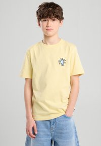 Keltainen puuvillapaita, jossa on pyöreä kaula-aukko ja lyhyet hihat, ja jossa on sininen brodeerattu kuvasto vasemmassa rintamuksessa.