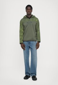 Sweatshirt verde com design gráfico tonal, gola com zíper e logótipo no peito, combinado com jeans azul claro e sapatos pretos.