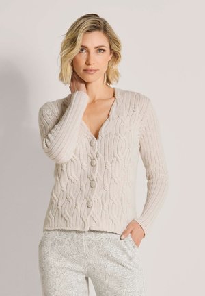Blonde vrouw met kort haar draagt een crèmekleurige kabelgebreide cardigan en lichte broek met paisley-patroon, poserend met één hand op haar nek.