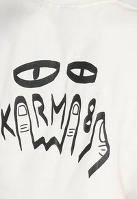 Weißer Stoff mit einem schwarzen Grafikdesign, das zwei Augen und Hände zeigt und durch kräftige Linien sowie eine minimalistische Ästhetik besticht. Der Text lautet "KARMA."