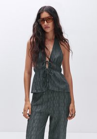Tamnozelena halter top s vezanjem sprijeda i teksturiranom tkaninom, uparen s usklađenim teksturiranim hlačama, koje imaju opušteni kroj.