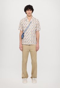 Kurzarmhemd mit hellem cremefarbenem Hintergrund und kleinen, bunten Fahrzeugmustern; kombiniert mit beigen Hosen und weißen Sneakers.