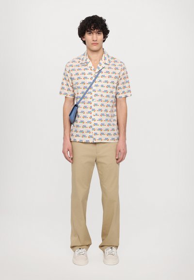 Chemise à manches courtes avec un fond crème clair et de petits motifs de véhicules colorés ; associée à un pantalon beige et des baskets blanches.