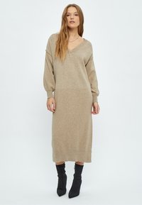 Bež haljina od pletiva s V-izrezom, dugim rukavima i slobodnim krojem, koja pada do sredine lista. Uparena s crnim čizmama na visoku petu s oštrim vrhom.