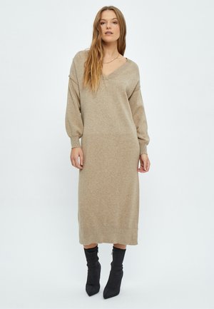 Beige strikket kjole med V-udskæring, lange ærmer og en løs pasform, der falder til midt på læggen. Sæt sammen med sorte spidse hæle støvler.