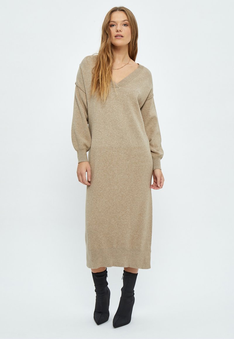 Bež haljina od pletiva s V-izrezom, dugim rukavima i slobodnim krojem, koja pada do sredine lista. Uparena s crnim čizmama na visoku petu s oštrim vrhom.