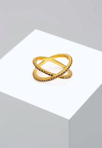 Bague de couleur dorée avec un design entrelacé, agrémentée de petites pierres noires le long de la bande, reposant sur un piédestal blanc.