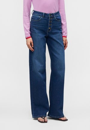 Frau, die dunkelblaue, hoch taillierte, weit geschnittene Jeans mit fünf goldenen Knöpfen und ein pinkfarbenes Langarmshirt trägt, steht vor einem schlichten Hintergrund.
