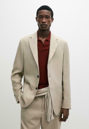Giovane uomo con indosso un completo beige, un maglione legato in vita e una camicia rossa con colletto, in piedi davanti a uno sfondo neutro.