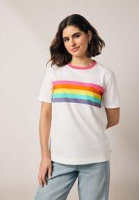 Fehér póló, amelyen vízszintes csíkok találhatók rózsaszín, narancssárga, sárga, zöld és lila színekben, rózsaszín kerek nyakkivágással és rövid ujjakkal.