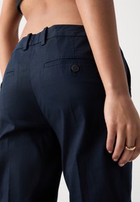 Personne portant un pantalon bleu marine foncé avec des poches arrière boutonnées, montrant le ventre nu et une main avec une bague en or reposant sur le côté.
