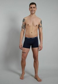 Blue boxer shorts made of soft fabric with a plain design. Features a branded waistband and a mid-thigh fit, model stands barefoot.