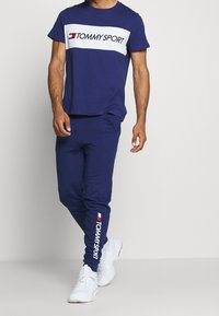 Mörkblå träningskläder bestående av en kortärmad T-shirt med en vit "TOMMY SPORT"-logotyp och matchande sweatpants med logotypdetaljer vid vristen.