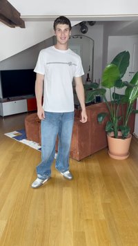 Balta marškinėliai su grafika, mėlynos plačios kojų džinsai su karoliukų apvadu ir sidabriniai batai. Fone matyti ruda sofa ir gėlės.
