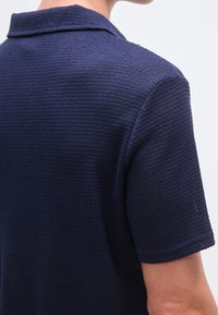 Marineblå poloskjorte med teksturert stoff, med krage og korte ermer, som viser en nærbilde av bakre design og sømdetaljer.