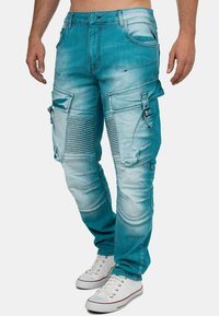 Un pantalon cargo bleu clair en denim, doté de multiples poches, d'accents texturés et d'un design de jambe fuselée. Associé à des baskets blanches.
