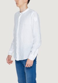 Camicia bianca in lino con colletto tondo e chiusura a bottoni, con maniche lunghe e tessuto strutturato. Pantaloni blu completano il look.