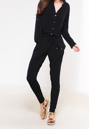 Kvinde iført en langærmet sort jumpsuit med gyldne knapper og snøre i taljen, kombineret med gyldne platformsandaler, stående på en lys baggrund.