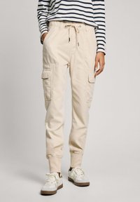 Beige cargo pants with elastic waistband, side pockets, and elastic cuffs. Paired with white sneakers featuring light brown accents.