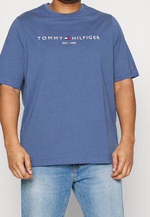 Mand iført en blå Tommy Hilfiger t-shirt med logo og "EST. 1985" tekst, kombineret med lyseblå jeans, stående mod en ensfarvet baggrund.