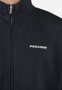 Crna jakna na zip s visokim ovratnikom, izrađena od teksturiranog materijala. Sadrži bijeli logo "PEGADOR" na lijevoj strani prsnog područja.