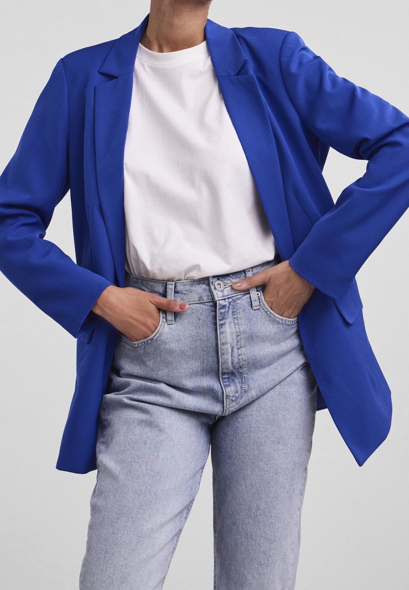 Personne portant un blazer bleu par-dessus un t-shirt blanc rentré dans un jean taille haute bleu clair, les mains dans les poches.