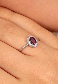Silberner Ring mit einem ovalen Rubin in der Mitte, umgeben von einem Halo aus kleinen klaren Steinen, auf einem glatten Band.