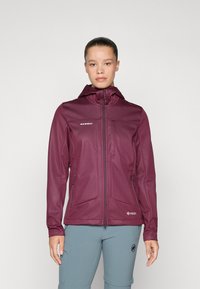 Woman standing facing forward wearing a zipped maroon hooded jacket and light blue pants with her hair tied back.