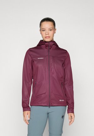 Woman standing facing forward wearing a zipped maroon hooded jacket and light blue pants with her hair tied back.