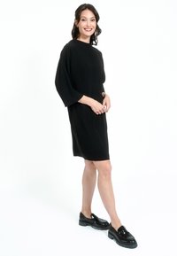 Črna obleka do kolen z okroglo izrezom in rokavi do tri četrtine, izdelana iz mehkega blaga. Model nosi črne čevlje s širokim petami.