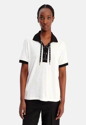 Donna che indossa una polo bianca a maniche corte con colletto nero, bordi delle maniche neri e dettaglio con lacci neri, abbinata a pantaloni neri, in piedi.