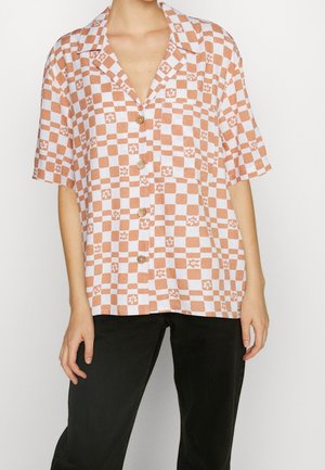 Chemise à manches courtes boutonnée avec un motif à carreaux marron et blanc agrémenté de petits accents floraux, portée avec un pantalon noir.