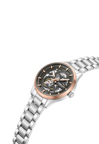 Sidabrinis nerūdijančio plieno laikrodis su juodu skeletiniu ciferblatu, rožinės auksinės spalvos akcentais ir sujungtu apyrankės dizainu. Yra valandų žymekliai ir karūnėlė.