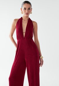 Mono burdeos tipo halter con escote en V profundo, piernas anchas y cintura ajustada. Tela suave con textura sutil. Pendientes dorados gruesos.