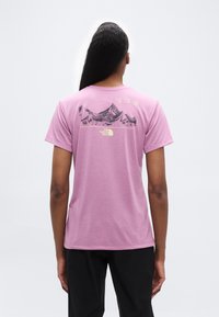 Άτομο με μακριά μαλλιά που φοράει μωβ μπλουζάκι με μαύρο γραφικό βουνού και ανοιχτόκκινο κίτρινο κείμενο και λογότυπο στην πλάτη, πάνω σε γκρι φόντο.