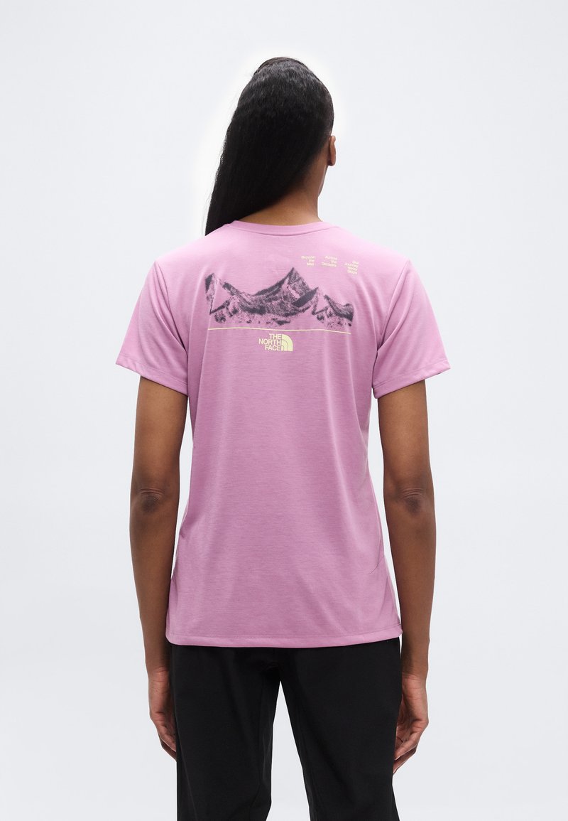 Άτομο με μακριά μαλλιά που φοράει μωβ μπλουζάκι με μαύρο γραφικό βουνού και ανοιχτόκκινο κίτρινο κείμενο και λογότυπο στην πλάτη, πάνω σε γκρι φόντο.