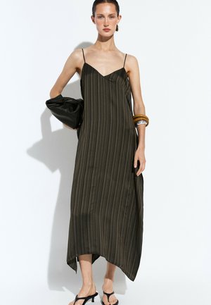 Donna che indossa un abito midi nero a righe senza maniche con spalline sottili, tiene in mano una pochette nera e indossa grandi braccialetti al polso.