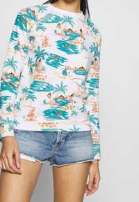 Sweat-shirt en coton à manches longues avec un motif coloré de plage et de palmiers, comprenant des tons de bleu, vert et rose, assorti à un short en denim.