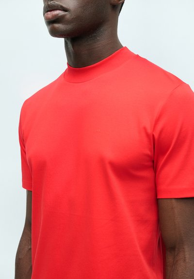 Κοντινό πλάνο ενός ατόμου που φοράει ένα απλό κόκκινο κοντομάνικο T-shirt με ψεύτικο λαιμό σε ανοιχτό φόντο.