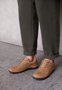 Chaussures en daim beige avec bout rond, deux œillets et coutures contrastées. Portées avec un pantalon vert olive retroussé. Semelle en caoutchouc texturée visible.