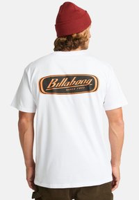 Hvid T-shirt med et sort og orange "Billabong" logo på ryggen, med et enkelt crew neck design og korte ærmer. Fremstillet af bomuld.